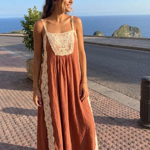 May include: A floor-length, rust-colored sleeveless dress featuring a square neckline with cream-colored lace detailing on the bodice and vertical lace panels running down the sides. The fabric has a textured, crinkled appearance throughout.