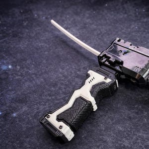 Puede incluir: Un lanzador de juguetes en blanco y negro con un cord&oacute;n blanco. El lanzador tiene un agarre texturizado y una pieza en forma de cruz en el extremo. El juguete est&aacute; sobre un fondo oscuro y moteado.