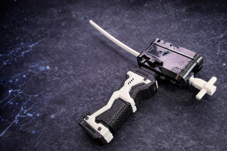 Puede incluir: Un lanzador de juguetes en blanco y negro con un cord&oacute;n blanco. El lanzador tiene un agarre texturizado y una pieza en forma de cruz en el extremo. El juguete est&aacute; sobre un fondo oscuro y moteado.