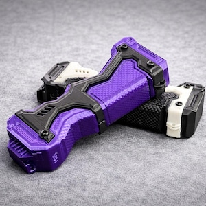 Puede incluir: Objetos impresos en tres dimensiones en p&uacute;rpura, negro, blanco y gris. El objeto principal es un dispositivo p&uacute;rpura y negro con una superficie texturizada. Otro objeto es blanco y negro. Los objetos est&aacute;n sobre una superficie gris.