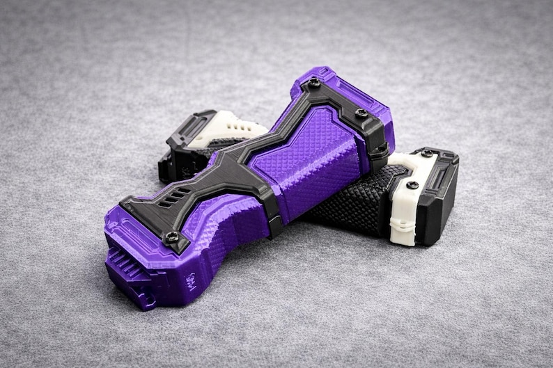 Puede incluir: Objetos impresos en tres dimensiones en p&uacute;rpura, negro, blanco y gris. El objeto principal es un dispositivo p&uacute;rpura y negro con una superficie texturizada. Otro objeto es blanco y negro. Los objetos est&aacute;n sobre una superficie gris.