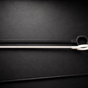 May include: Two cable ties, one black and one white, are displayed against a dark background. The black tie has a ridged surface and a loop at one end. The white tie is smooth with a loop at the end. These are useful for organizing cables.