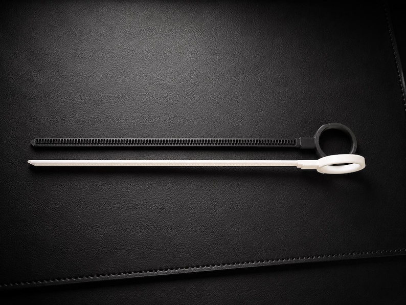 May include: Two cable ties, one black and one white, are displayed against a dark background. The black tie has a ridged surface and a loop at one end. The white tie is smooth with a loop at the end. These are useful for organizing cables.