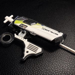 May include: A white, black, and yellow Beyblade X launcher and accessories are displayed on a black surface. The launcher is rectangular with the text "BEYBLADE X" on the side. A white handle and a black and silver circular piece are also present.