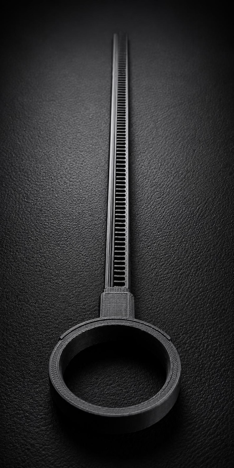 May include: A black, 3D-printed tool with a long, ridged handle and a circular base. The handle has a series of parallel lines running along its length. The tool is set against a dark, textured background.