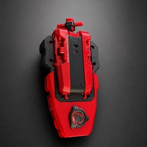 May include: A red and black Beyblade X String Launcher toy with a T-shaped handle at the top. The device features a central black panel with the text BEYBLADE X STRING LAUNCHER printed in white and a circular black emblem with red graphics at the base.
