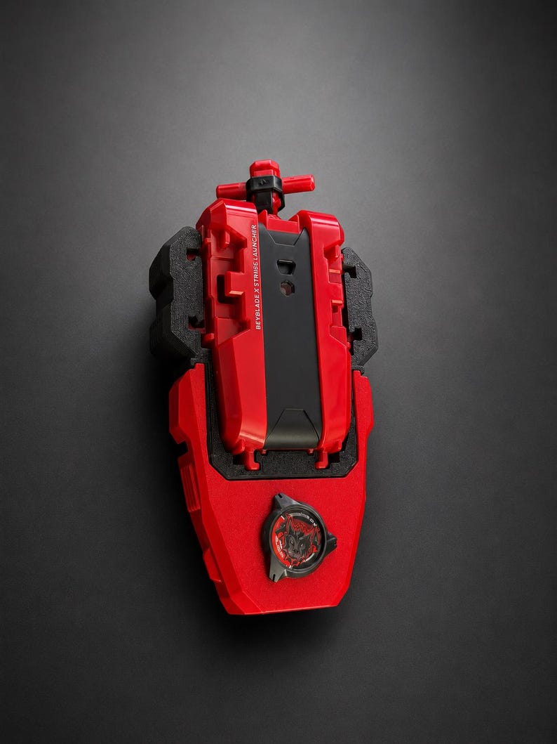 May include: A red and black Beyblade X String Launcher toy with a T-shaped handle at the top. The device features a central black panel with the text BEYBLADE X STRING LAUNCHER printed in white and a circular black emblem with red graphics at the base.