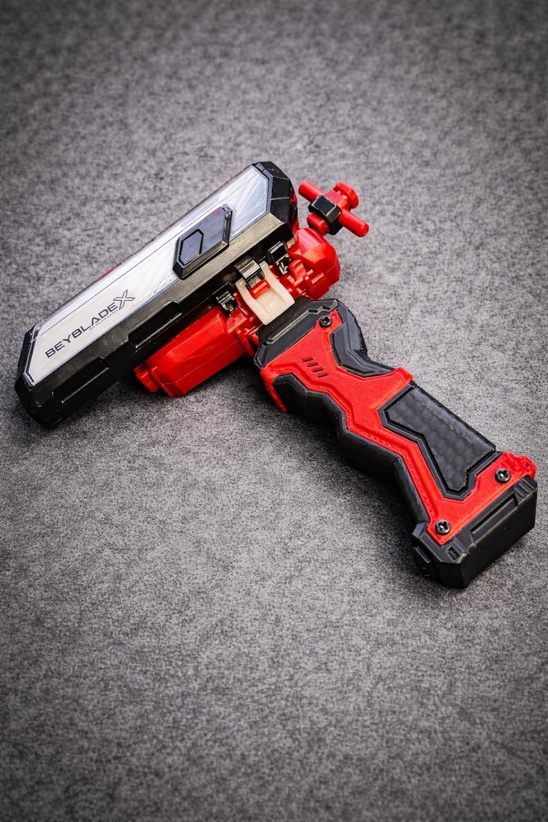 Puede incluir: Un lanzador Beyblade X rojo y negro con una secci&oacute;n superior plateada y negra. El lanzador tiene un agarre texturizado y el texto "BEYBLADEX" es visible en el lateral. El lanzador est&aacute; dise&ntilde;ado para lanzar peonzas Beyblade.