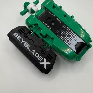 May include: A green and black Beyblade X string launcher with a white handle grip. The device features the text BEYBLADE X on the black section and STRING LAUNCHER on the green body, with a dotted pattern and a small white cross-shaped component.