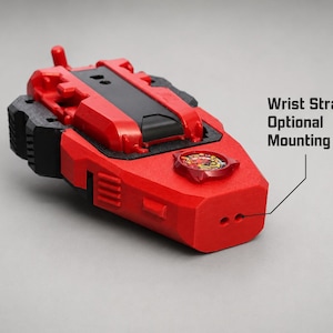 May include: A red and black plastic device featuring a central black panel, a circular emblem with a graphic design, and a front-facing section with two small holes labelled as a Wrist Strap Optional Mounting Point for attachment purposes.