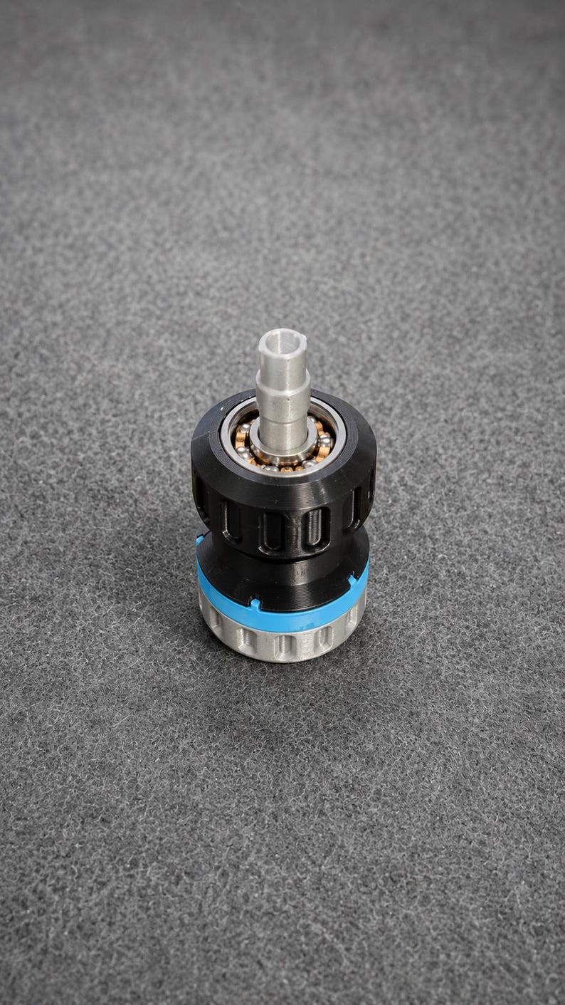 May include: A mechanical component featuring a central silver metal shaft extending from a black cylindrical housing. The assembly includes a visible ball bearing ring, a bright blue accent ring, and a silver base with vertical grooves.