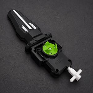Puede incluir: Lanzador de juguete negro y verde con un cord&oacute;n blanco. El lanzador tiene un mango negro con detalles plateados y un componente circular verde. El juguete est&aacute; dise&ntilde;ado para lanzar una peonza.