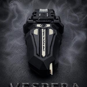 Puede incluir: Un dispositivo negro y geom&eacute;trico con detalles blancos y la palabra "VESPERA" en la parte inferior. El dispositivo tiene un dise&ntilde;o complejo con una superficie texturizada y una peque&ntilde;a palanca blanca. El fondo es oscuro con un efecto ahumado.