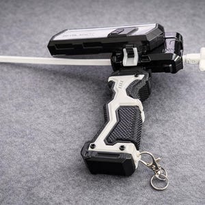 Puede incluir: Un lanzador de juguete en blanco y negro con un cord&oacute;n blanco y un llavero plateado. El lanzador tiene un agarre texturizado y una pieza blanca en forma de cruz en el extremo. El juguete est&aacute; dise&ntilde;ado para lanzar una peonza.