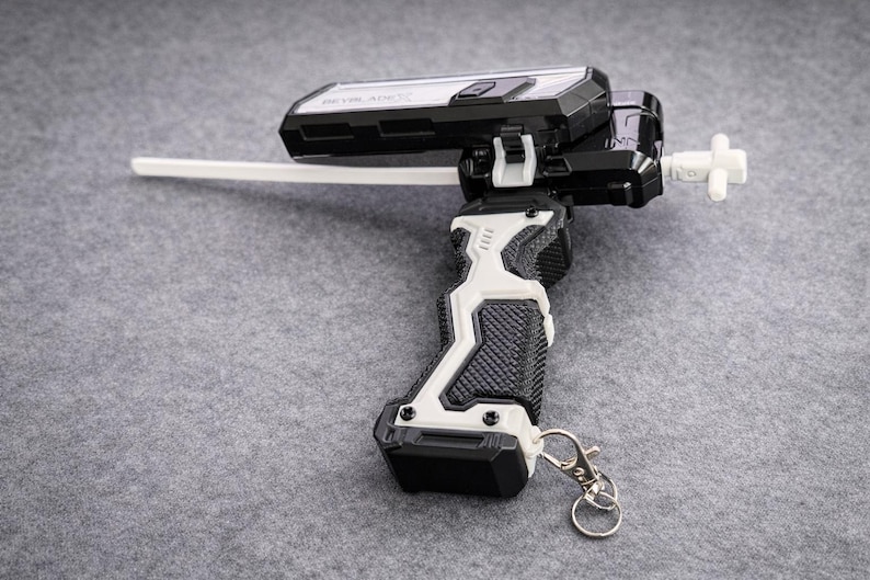 Puede incluir: Un lanzador de juguete en blanco y negro con un cord&oacute;n blanco y un llavero plateado. El lanzador tiene un agarre texturizado y una pieza blanca en forma de cruz en el extremo. El juguete est&aacute; dise&ntilde;ado para lanzar una peonza.