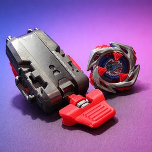 May include: A black and red Beyblade X string launcher and a red, silver, and blue Beyblade top are displayed on a purple and blue gradient background. The launcher has the text "Beyblade X String Launcher".