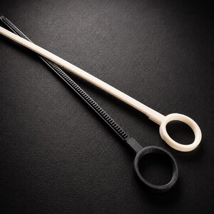 May include: Two plastic loop tools, one black and one off-white, are angled across a dark surface. Each tool has a long, thin handle with a ridged section and a circular loop at the end. The tools appear to be new.