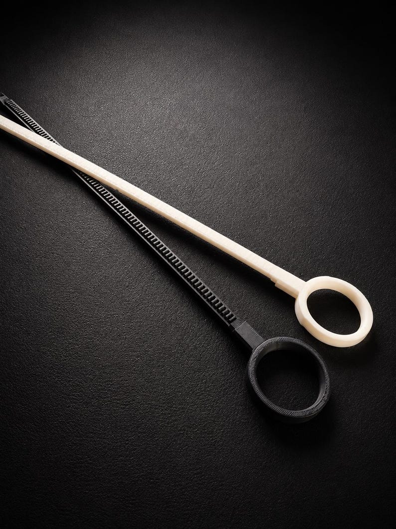 May include: Two plastic loop tools, one black and one off-white, are angled across a dark surface. Each tool has a long, thin handle with a ridged section and a circular loop at the end. The tools appear to be new.
