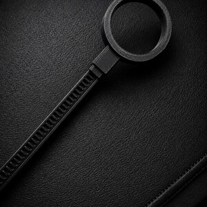 May include: A black, circular tool with a handle, resembling a magnifying glass or a specialized fastener. The handle has a ridged design, and the circular end is smooth. The object is set against a textured, dark background.