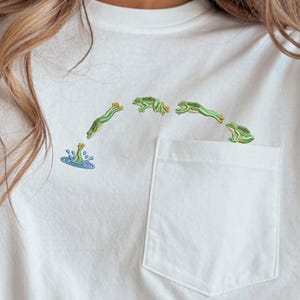 Puede incluir: Una camiseta blanca con un bolsillo en el pecho y un bordado de una rana verde saltando en arco desde el bolsillo hacia una pequeña salpicadura de agua azul. El diseño muestra a la rana en cuatro posiciones distintas durante el salto.