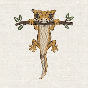 Può includere: Un disegno da ricamo che raffigura un geco crestato color marrone chiaro e scuro appeso a un ramo d'albero orizzontale marrone scuro. Il geco ha grandi occhi scuri, un ampio sorriso e piccole foglie verdi alle estremità del ramo legnoso.