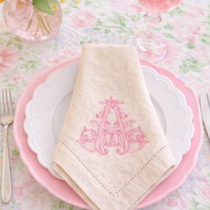 May include: A beige linen napkin featuring a decorative pink embroidered letter A in a scrollwork font. The napkin is folded into a triangle and placed on a white scalloped plate, which sits atop a larger pink plate with a scalloped edge.