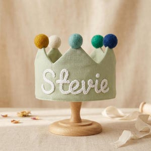 May include: A sage green fabric crown featuring the name Stevie embroidered in white script lettering. The crown is adorned with five colorful felt pom-poms in mustard yellow, cream, teal, green, and navy blue, positioned on each pointed tip.