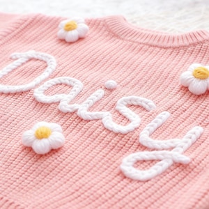 Pode incluir: Um close-up de um suéter de malha rosa com o nome "Daisy" bordado em branco. O suéter é adornado com aplicações de margaridas brancas com centros amarelos, criando um design floral. O tecido de malha tem uma textura canelada.