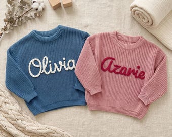 Peut inclure: Deux pulls en tricot à manches longues avec encolures rondes côtelées. Un pull est bleu avec le prénom Olivia brodé en écriture blanche. L'autre pull est rose avec le prénom Azarie brodé en écriture rouge foncé.