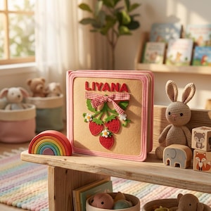 May include: A square, beige felt book with a pink border, featuring the name LIYANA in red letters above a red and white gingham bow. Below the bow are three red strawberry appliqués with green leaves and small white flowers.