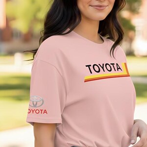 May include: A light pink t-shirt with the word "TOYOTA" in black letters and a yellow and red stripe design. The Toyota logo is on the left sleeve. The shirt is made of a soft material.
