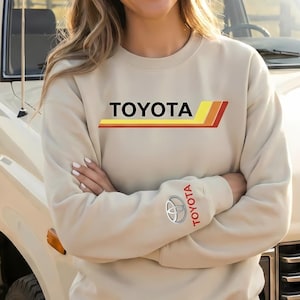 May include: Beige sweatshirt with the word "TOYOTA" in black block letters and a red, yellow, and orange stripe design. The Toyota logo and the word "TOYOTA" are printed on the left sleeve. The sweatshirt is worn by a person standing next to a vintage Toyota SUV.