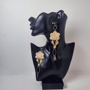 Puede incluir: Un par de pendientes colgantes dorados y blancos con un diseño en forma de nube. Los pendientes cuelgan de una cabeza de maniquí negra.