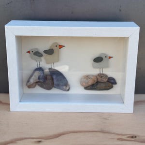 May include: A framed artwork depicting three birds crafted from glass and pebbles. The birds have grey bodies, black wing details, and orange beaks. They are positioned on rocks within a white wooden frame, evoking a seaside theme.