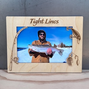 Peut inclure: Cadre photo en bois avec une photo d'une personne tenant un gros poisson. Le cadre présente des motifs sur le thème de la pêche découpés au laser et les mots "Tight Lines" en haut. La photo montre un paysage enneigé et un ciel bleu.