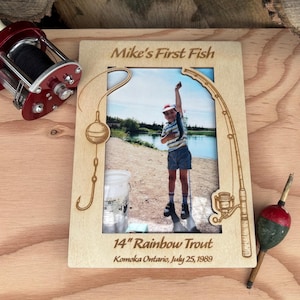 Puede incluir: Un marco de fotos de madera con el texto "Mike's First Fish" y una foto de un niño sosteniendo un pez. El marco incluye el texto "14" Rainbow Trout" y "Komoka Ontario, July 25, 1989". También se ven un carrete de pesca rojo y un flotador.