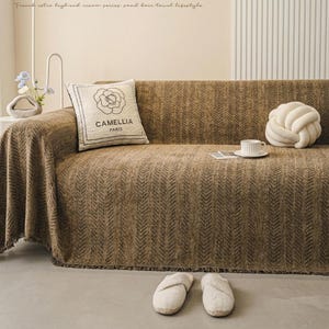 Puede incluir: Un sofá marrón con una manta estampada y un cojín decorativo con el texto "CAMELLIA PARIS". Una almohada blanca en forma de nudo y una taza y platillo también están en el sofá. Dos pares de zapatillas blancas están en el suelo.