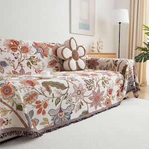 May include: A sofa adorned with a floral patterned cover and a flower-shaped cushion. The cover displays a cream backdrop with orange, blue, and green floral patterns. A white teacup rests on the sofa. The sofa sits on a white rug.