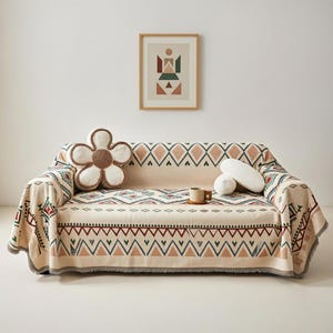 May include: A sofa draped with a beige patterned throw, adorned with geometric shapes in red, green, and blue. A flower-shaped cushion and two white pillows are on the sofa. A framed abstract artwork hangs above.