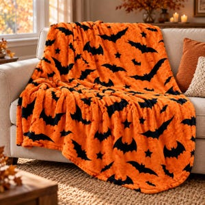 Könnte beinhalten: Eine flauschige orangefarbene Wohndecke mit einem sich wiederholenden Muster aus schwarzen Fledermäusen und kleinen Sternen. Der Stoff hat eine weiche, strukturierte Optik und liegt über einem Sofa, wobei er Sitz und Rückenlehne bedeckt.