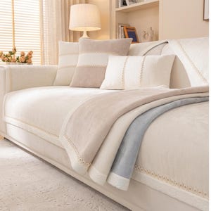 Puede incluir: Un conjunto de fundas para sofá en color beige con una textura suave y afelpada, adornado con detalles de encaje color crema. Incluye una funda rectangular para el asiento, cojines cuadrados y rectangulares a juego y una manta decorativa.