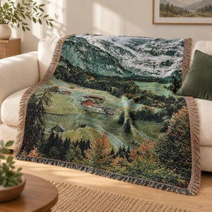 May include: A woven throw blanket featuring a detailed landscape scene of snow-capped mountains, green rolling hills, and small houses nestled among dense pine forests. The blanket has a fringed edge and a rich color palette of green, brown, and white.