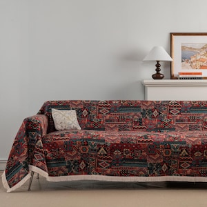 Może przedstawiać: Sofa przykryta narzutą w stylu patchwork. Narzuta ma mieszankę czerwonych, niebieskich i beżowych geometrycznych wzorów. Biała frędzel i dekoracyjna poduszka dodają przytulnej estetyki. Lampa i oprawione dzieło sztuki są widoczne w tle.