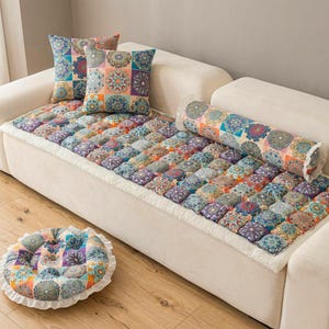 Puede incluir: Un sofá blanco con cojines estampados y una funda de asiento acolchada. Los cojines y la funda presentan un diseño de patchwork con tonos de azul, naranja, morado y turquesa. También hay un cojín redondo para el suelo con un borde con volantes.