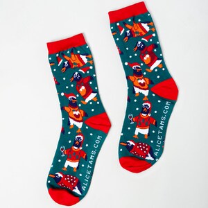 May include: A pair of festive socks with a teal background, featuring a pattern of penguins wearing Santa hats and sweaters. The socks have red heels, toes, and cuffs. The text "ALICETAMS.COM" is printed on the side of each sock.