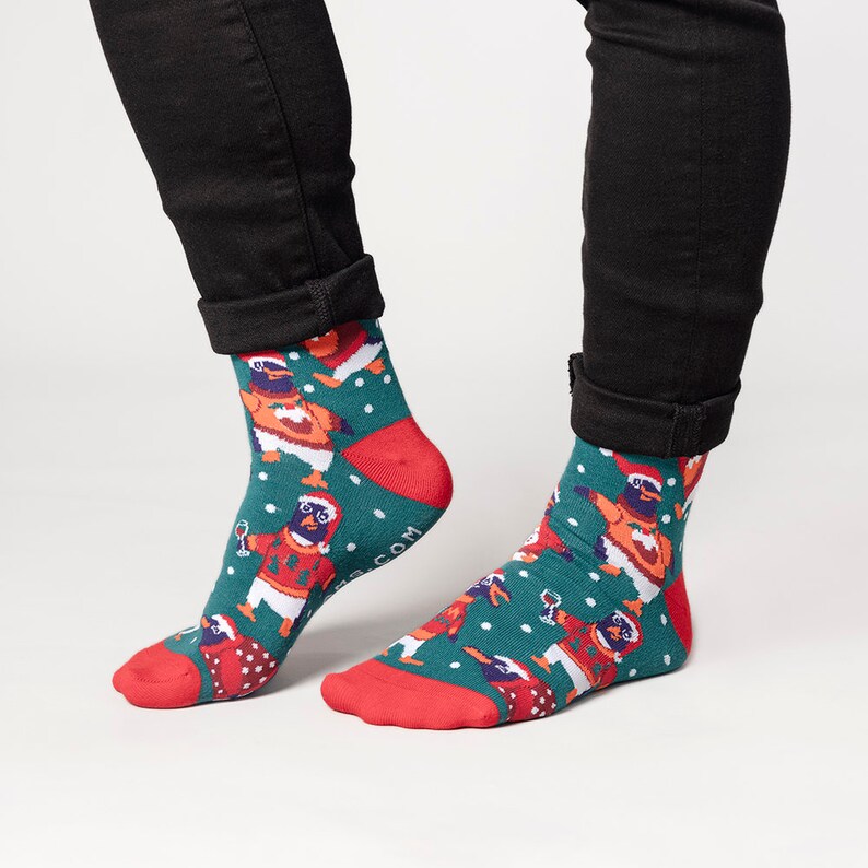 May include: A pair of festive socks with a teal background and a pattern of penguins wearing Santa hats and sweaters. The socks have red heels and toes, and white snowflakes are scattered across the design.