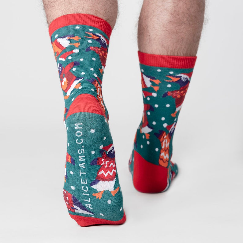 May include: A pair of green socks with a festive penguin pattern. The socks have red cuffs, heels, and toes. The design includes penguins wearing Santa hats and scarves, with the text ALICETAMS.COM printed vertically on the side.
