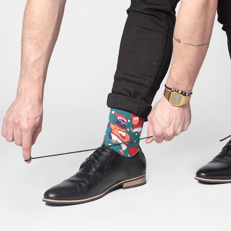 May include: A person tying the laces of a black leather dress shoe. The person is wearing a pair of teal socks with a cartoon design featuring a penguin in a Santa hat. The person is also wearing a gold watch and a beaded bracelet.