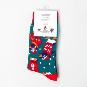 May include: A pair of teal socks with a festive penguin pattern in red, white, and orange. The socks have red cuffs and toes, and a white tag with care instructions and the sock's material composition. The socks are made of 80% combed cotton, 17% nylon, 2% spandex, and 100% kind love.
