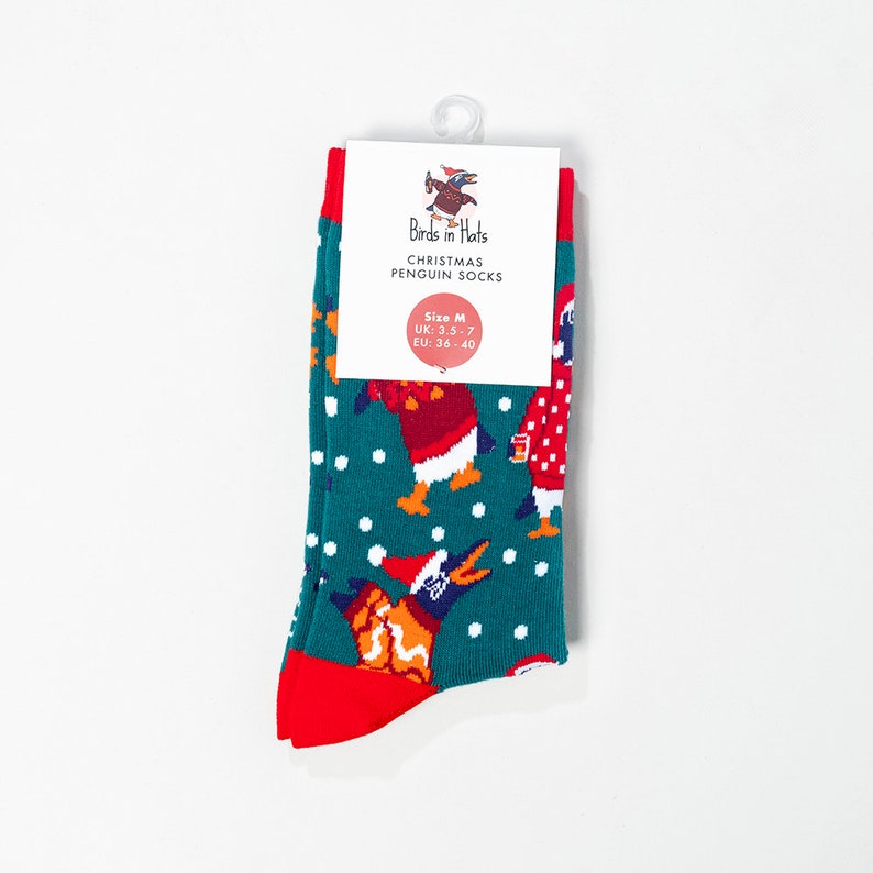 May include: A pair of Christmas penguin socks in teal, red, and white. The socks feature a repeating pattern of penguins wearing Santa hats and falling snowflakes. The socks have red cuffs and toes. The label reads "Birds in Hats Christmas Penguin Socks."